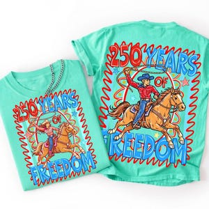Puede incluir: Una camiseta verde menta con un estampado gr&aacute;fico de una persona montando a caballo con un lazo. El texto dice 250 YEARS OF FREEDOM en rojo y azul. El dise&ntilde;o incluye un borde ondulado rojo, estrellas y un caballo marr&oacute;n con un jinete.