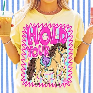 May include: A pale yellow t-shirt featuring a graphic print with the text "HOLD YOUR" in bold pink letters above an illustration of a tan horse with a purple saddle. The design is enclosed in a bright pink squiggly border with small stars.