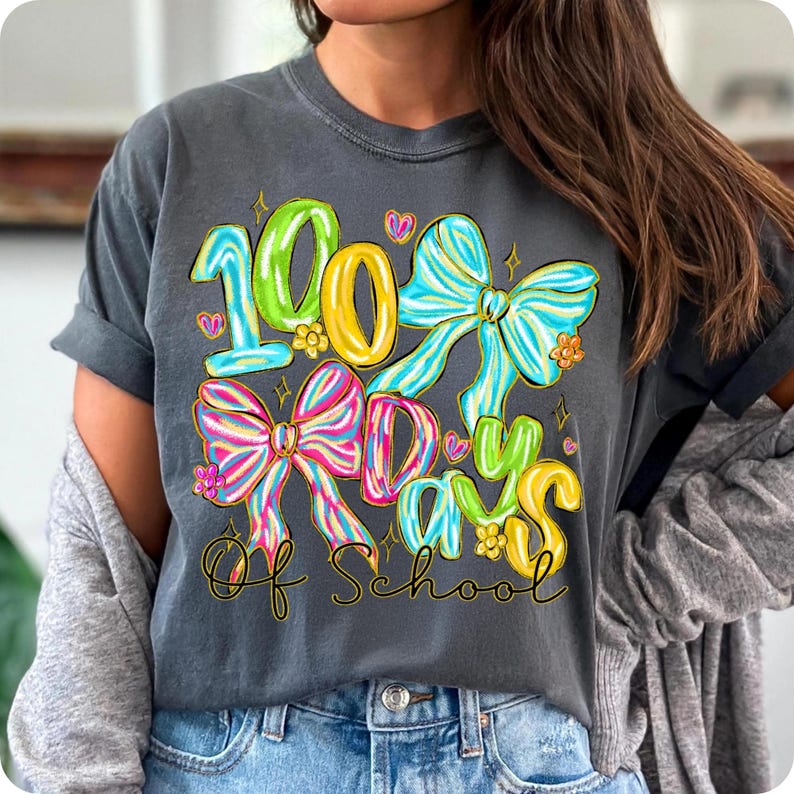 Op de afbeelding: Donkergrijs t-shirt met een kleurrijke afbeelding ter viering van 100 schooldagen. Het ontwerp toont de woorden "100 Days Of School" in heldere kleuren, met decoratieve strikken en harten. Een casual, comfortabel t-shirt.