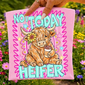 May include: Pink t-shirt with a cartoon illustration of a Highland cow holding a coffee cup, wearing a sunflower, and the text "NOT TODAY HEIFER" in turquoise. The design is surrounded by a pink wavy border and stars.