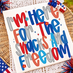 May include: A white t-shirt featuring the text I'M HERE FOR THE SNACKS AND FREEDOM in red, blue, and grey bubble letters. The design includes small flower and star accents in red, blue, and yellow, with a patriotic hair clip placed at the top.