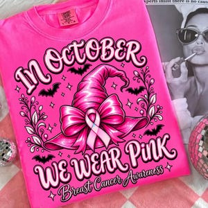 May include: A bright pink t-shirt featuring a graphic of a witch hat adorned with a pink ribbon. The text reads "IN OCTOBER WE WEAR PINK" and "Breast Cancer Awareness" with small bat and star illustrations surrounding the central design.