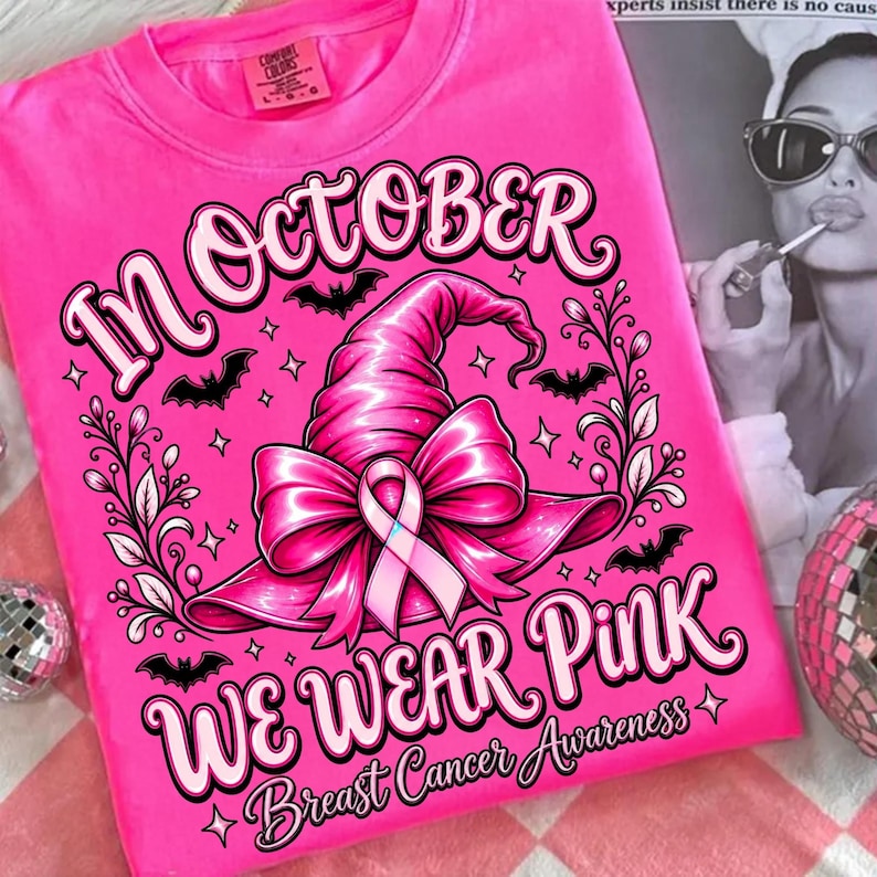 May include: A bright pink t-shirt featuring a graphic of a witch hat adorned with a pink ribbon. The text reads "IN OCTOBER WE WEAR PINK" and "Breast Cancer Awareness" with small bat and star illustrations surrounding the central design.