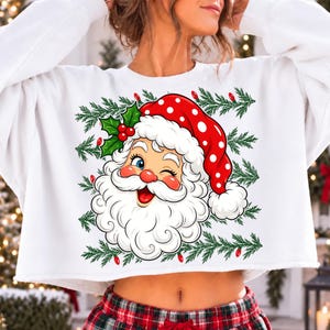May include: A white cropped sweatshirt featuring a graphic of a winking Santa Claus wearing a red hat with white polka dots. The design is framed by green pine branches and small red Christmas lights, creating a festive holiday-themed garment.