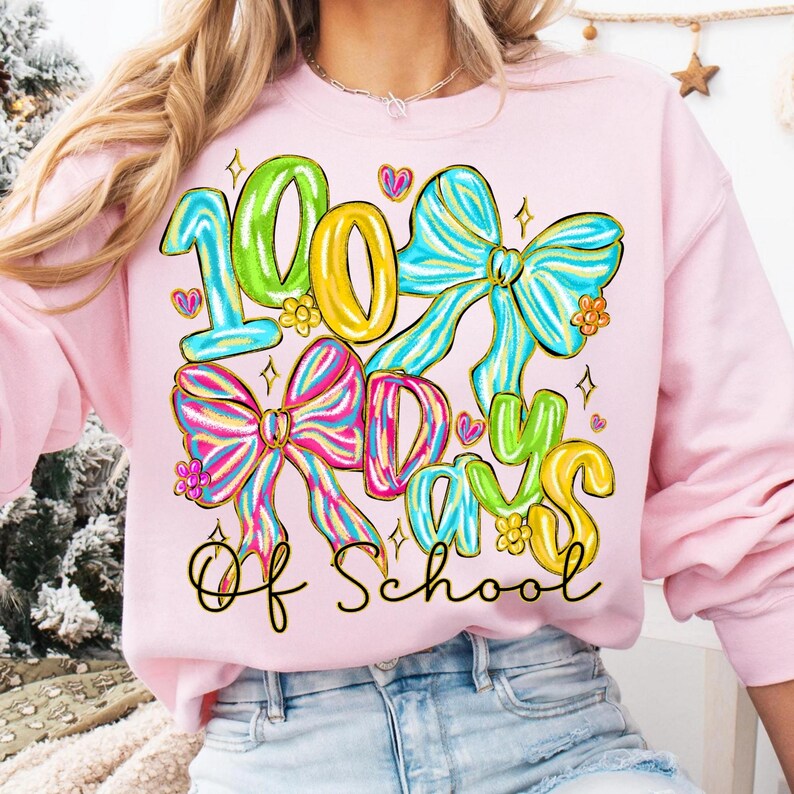 Op de afbeelding: Een lichtroze sweatshirt met de tekst "100 Days Of School" in kleurrijke, speelse letters. Het ontwerp bevat grote, decoratieve strikken in groen, geel, blauw en roze, met bloemaccenten en hartjes.