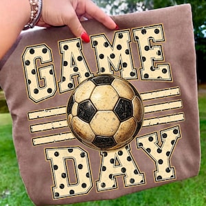 May include: A brown t-shirt featuring a graphic print of a soccer ball centered between the words GAME and DAY. The text is written in a large, blocky, cream-colored font with black polka dots, and the soccer ball has a distressed, vintage texture.