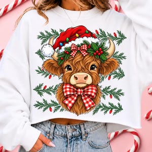 Puede incluir: Una sudadera blanca de manga larga con el gr&aacute;fico de una vaca Highland marr&oacute;n que lleva un gorro de Pap&aacute; Noel rojo, una corona de acebo verde y un lazo de cuadros rojos y blancos. El fondo incluye ramas de pino verdes y luces rojas.