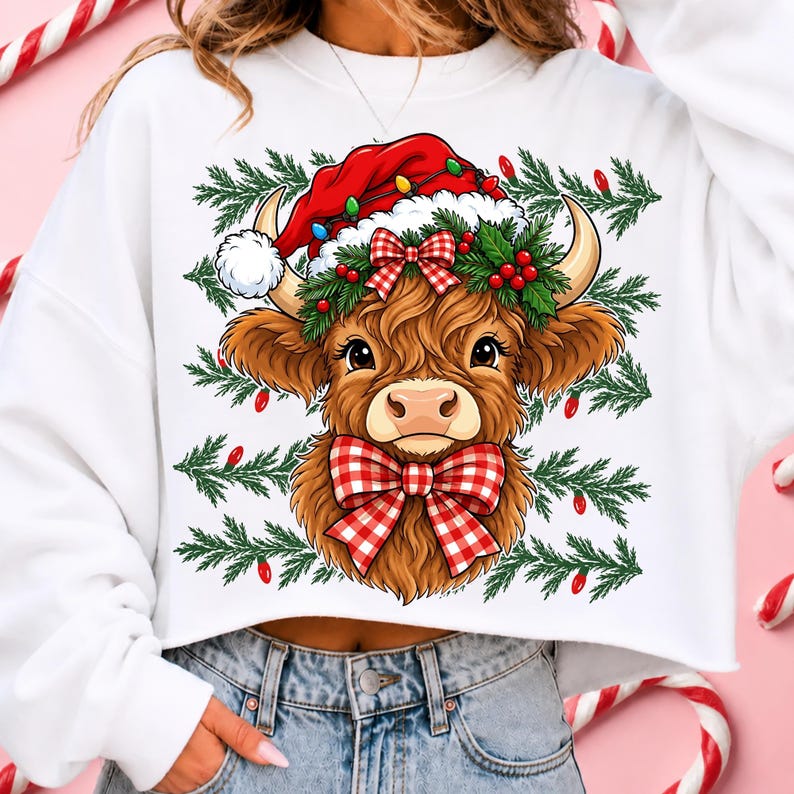 Puede incluir: Una sudadera blanca de manga larga con el gr&aacute;fico de una vaca Highland marr&oacute;n que lleva un gorro de Pap&aacute; Noel rojo, una corona de acebo verde y un lazo de cuadros rojos y blancos. El fondo incluye ramas de pino verdes y luces rojas.