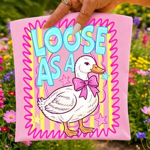 K&ouml;nnte beinhalten: Ein hellrosa Stoffartikel mit einer wei&szlig;en Entenillustration, die eine leuchtend rosa Schleife um den Hals tr&auml;gt. Der Text LOOSE AS A ist in gro&szlig;en hellblauen Buchstaben &uuml;ber der Ente gedruckt. Das Design enth&auml;lt gelbe vertikale Streifen und rosa Sterne.