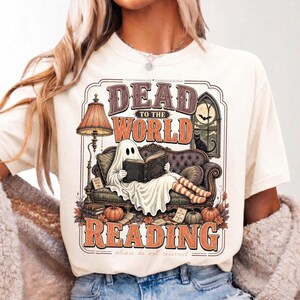 May include: A cream-colored t-shirt featuring a graphic of a ghost sitting on a sofa reading a book. The text reads "DEAD TO THE WORLD READING" and "please do not resurrect". The illustration includes a lamp, a black cat, and pumpkins.