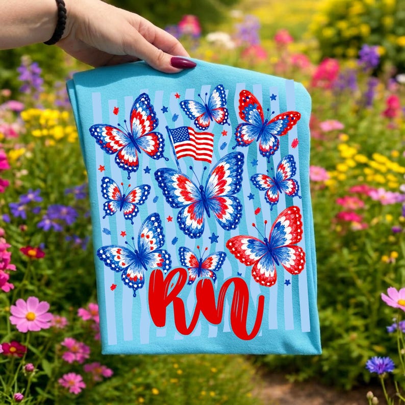May include: A light blue t-shirt featuring a pattern of butterflies with red, white, and blue wings, a small American flag, and the red cursive text RN. The design includes vertical light blue stripes and small red and blue stars throughout.