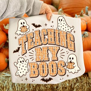 May include: A white t-shirt featuring a graphic print with the text "TEACHING MY BOOS" in an orange and white gingham pattern. The design includes four cartoon ghosts, some with polka dots, holding small pumpkins, and several small black bats.