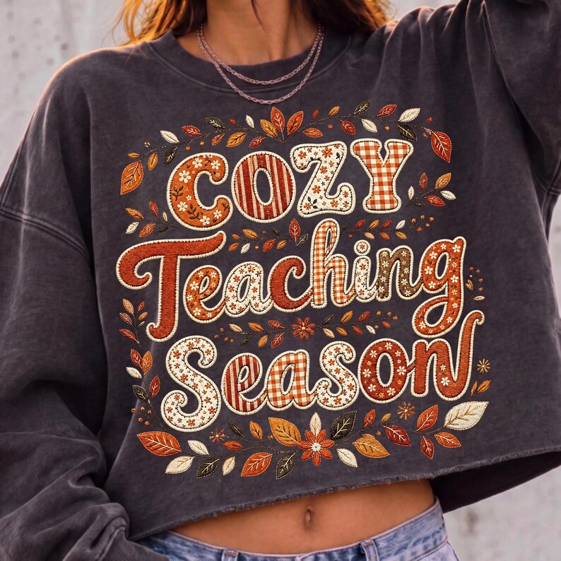 May include: A charcoal gray cropped sweatshirt featuring the text "COZY Teaching Season" in a decorative font. The letters are filled with orange, brown, and white patterns including gingham, stripes, and floral motifs, surrounded by autumn leaves.