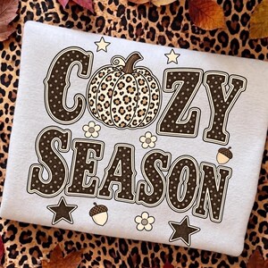 Może przedstawiać: Biała bluza z napisem COZY SEASON ciemnobrązowymi literami w białe kropki. Centralna grafika dyni ma wz&oacute;r w panterkę. Dodatkowe elementy graficzne to małe białe kwiatki, brązowe gwiazdki oraz brązowe żołędzie.