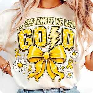 May include: A cream-colored t-shirt featuring a graphic with the text IN SEPTEMBER WE WEAR GOLD. The design includes a large yellow polka-dot word GOLD with a lightning bolt as the letter L, a large yellow bow, small yellow ribbons, and daisies.