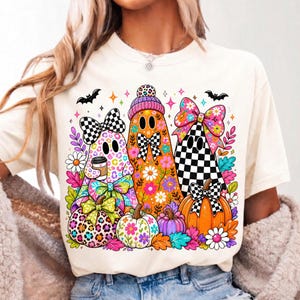 Puede incluir: Una camiseta de color crema con un gr&aacute;fico de tres fantasmas decorados con patrones coloridos, incluyendo estampado de leopardo, dise&ntilde;os florales y lazos a cuadros. Los fantasmas est&aacute;n rodeados de calabazas, flores y murci&eacute;lagos.