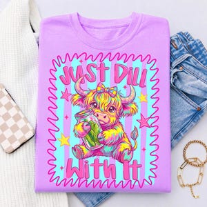Puede incluir: Una camiseta de color morado claro con un gr&aacute;fico colorido de una vaca Highland con un lazo, sosteniendo un frasco de cristal con pepinillos. El texto "JUST DILL WITH IT" est&aacute; impreso en rosa, rodeado por un borde ondulado rosa y estrellas.