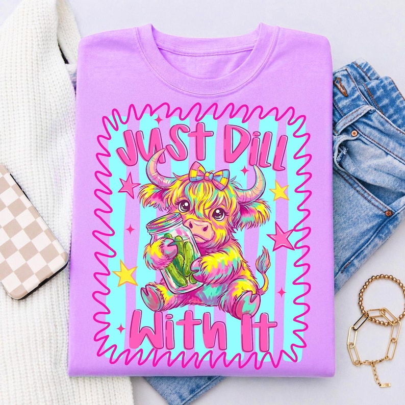 Puede incluir: Una camiseta de color morado claro con un gr&aacute;fico colorido de una vaca Highland con un lazo, sosteniendo un frasco de cristal con pepinillos. El texto "JUST DILL WITH IT" est&aacute; impreso en rosa, rodeado por un borde ondulado rosa y estrellas.