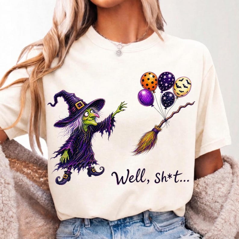 K&ouml;nnte beinhalten: Ein cremefarbenes T-Shirt mit einer Grafik einer gr&uuml;nh&auml;utigen Hexe in violettem Gewand und Hut, die nach einem Besen greift, an dem f&uuml;nf bunte Luftballons befestigt sind. Der Text "Well, Sh*t..." ist in schwarzer Schrift darunter gedruckt.