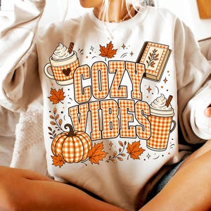 Puede incluir: Una sudadera color crema con un estampado gr&aacute;fico que incluye el texto COZY VIBES en un patr&oacute;n de cuadros naranja y blanco. El dise&ntilde;o presenta una calabaza, dos tazas con crema batida y un libro, todo acentuado con hojas de arce naranjas.