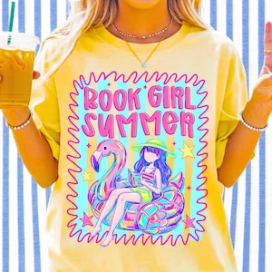 May include: A yellow t-shirt featuring a graphic print with the text BOOK GIRL SUMMER in pink. The design includes a person wearing a hat and reading a book while sitting on a pink flamingo pool float, surrounded by yellow stars and a blue striped background.
