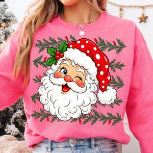May include: A bright pink crewneck sweatshirt featuring a large graphic of a winking Santa Claus wearing a red hat with white polka dots. The design includes green pine branches and small red light bulbs surrounding the central Santa illustration.