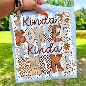Pode incluir: Uma t-shirt azul-clara com um design gr&aacute;fico contendo o texto Kinda BOUJEE Kinda BROKE. O design inclui padr&otilde;es xadrez castanhos e bege, pontos, pequenas estrelas, flores e um la&ccedil;o, dispostos num esquema de riscas verticais.