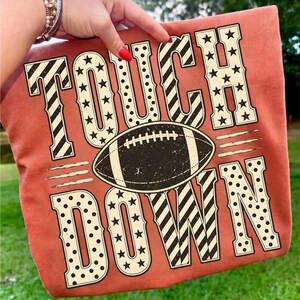 以下が含まれることがあります： 星と水玉模様の大きな装飾フォントで「TOUCH DOWN」という文字がプリントされた、バーントオレンジ色のTシャツ。文字の間には、白いレースが付いたダークブラウンのフットボールのグラフィックが中央に配置されており、かすれたような質感が施されています。