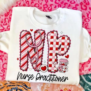 May include: White sweatshirt with the letters "NP" in red and pink, featuring a stethoscope and hearts. The letter "N" has red and white stripes, and the letter "P" has a red and white checkerboard pattern. The words "Nurse Practitioner" are written below.