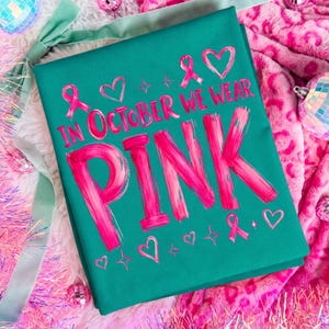 May include: A teal t-shirt featuring the bright pink text IN OCTOBER WE WEAR PINK, accented with pink awareness ribbons, heart shapes, and small four-pointed star graphics. The design is printed in a bold, brushstroke-style font.