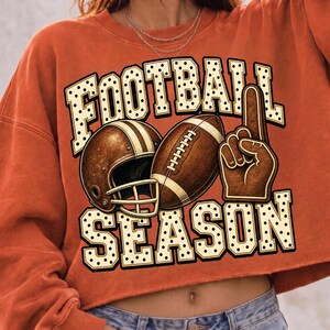 Puede incluir: Una sudadera naranja de manga larga con un estampado gr&aacute;fico que muestra el texto FOOTBALL SEASON en letras grandes de color crema con puntos negros. El dise&ntilde;o incluye un casco de f&uacute;tbol americano, un bal&oacute;n y un dedo de espuma marr&oacute;n.