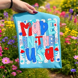 May include: A light blue t-shirt featuring the text MADE IN THE USA in a bold, multi-coloured font with red, white, and blue polka dot and checkered patterns. The design includes small firework and star graphics and two rocket icons.