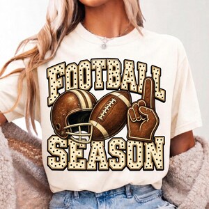 Puede incluir: Una camiseta de color crema con un estampado gr&aacute;fico que incluye el texto FOOTBALL SEASON en una fuente de puntos. El dise&ntilde;o muestra un casco de f&uacute;tbol americano marr&oacute;n, un bal&oacute;n de f&uacute;tbol y un dedo de espuma marr&oacute;n apuntando hacia arriba.