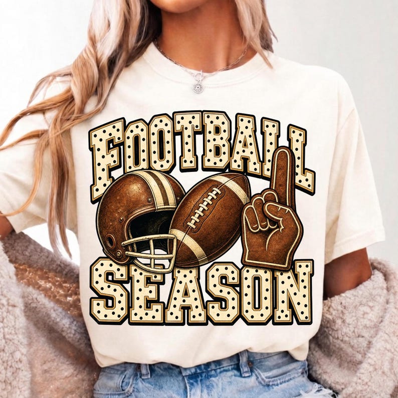 Puede incluir: Una camiseta de color crema con un estampado gr&aacute;fico que incluye el texto FOOTBALL SEASON en una fuente de puntos. El dise&ntilde;o muestra un casco de f&uacute;tbol americano marr&oacute;n, un bal&oacute;n de f&uacute;tbol y un dedo de espuma marr&oacute;n apuntando hacia arriba.