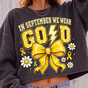 May include: A charcoal gray long-sleeve sweatshirt featuring a graphic print with the text IN SEPTEMBER WE WEAR GOLD. The design includes large yellow polka-dot letters, a yellow lightning bolt, a yellow bow, small yellow ribbons, and white daisies.