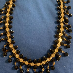 May include: A gold-toned necklace with black beads and clear, round rhinestones. The necklace has a delicate design with dangling bead clusters. The background is a solid blue.