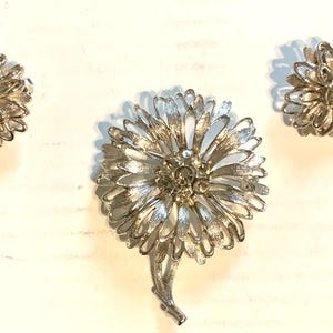 May include: A silver-toned floral jewelry set, including a brooch and matching clip-on earrings. The brooch features a large flower design with detailed petals and a stem. The earrings mirror the flower design. The set is likely vintage.