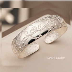 May include: A silver cuff bracelet with an engraved floral design. The bracelet features a detailed pattern of flowers and leaves, with a polished finish. The text "ELEVARY JEWELRY" is visible on the bottom right.