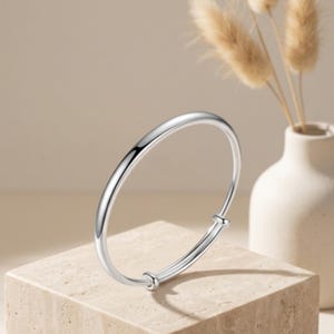 May include: A polished silver-tone bangle bracelet with a smooth, rounded surface. The bracelet features an adjustable design with two small, cylindrical sliding components that allow the diameter to be expanded or contracted for a secure fit.