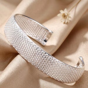 May include: A silver-colored cuff bracelet with a woven mesh design. The open-ended bracelet is displayed on a beige fabric, with a small, dried flower in the background. The bracelet has a shiny, metallic finish.