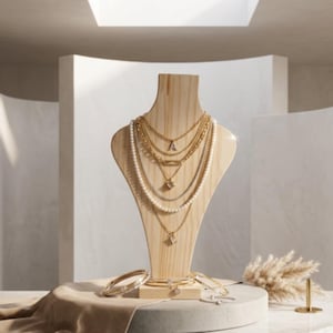 May include: A light-colored wooden jewelry display bust featuring multiple layered gold-tone necklaces, including one with a letter A pendant, and a strand of white pearls. Several gold-tone bracelets are arranged at the base of the display.