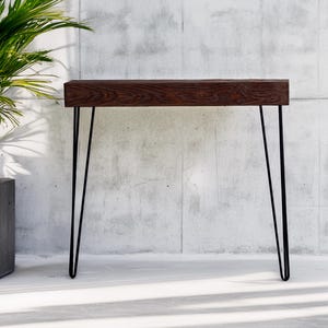 May include: A modern console table with a dark wood top and black hairpin legs. The table is approximately 48 inches long and 12 inches wide. A potted plant is to the left of the table against a concrete wall.