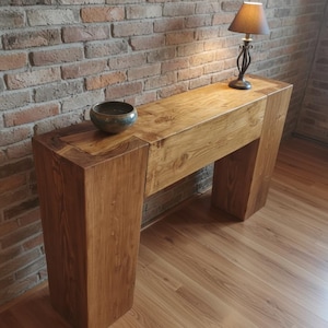 Może przedstawiać: Konsola z litego drewna z naturalnym wykończeniem. Stół ma prostokątny blat i dwie grube, skośne nogi. Na stole stoi mała lampa z brązowym kloszem i dekoracyjna miska. Tło stanowi ceglana ściana i drewniana podłoga.