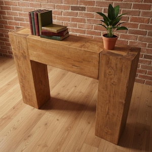 Pode incluir: Uma mesa de console de madeira marrom clara sólida com uma parte superior retangular e duas pernas quadradas grossas. A mesa é adornada com uma pequena planta em vaso e uma pilha de livros. O fundo apresenta uma parede de tijolos e piso de madeira.