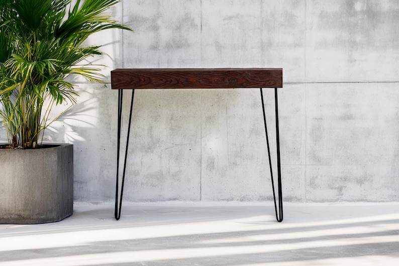 May include: A modern console table with a dark wood top and black hairpin legs. The table is approximately 48 inches long and 12 inches wide. A potted plant is to the left of the table against a concrete wall.