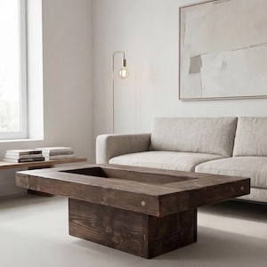 Op de afbeelding: Een donkerbruine rechthoekige salontafel met een verzonken midden. De tafel is gemaakt van dik, massief hout en heeft een natuurlijke houtnerf. De tafel staat in een woonkamer met een bank en een vloerlamp.