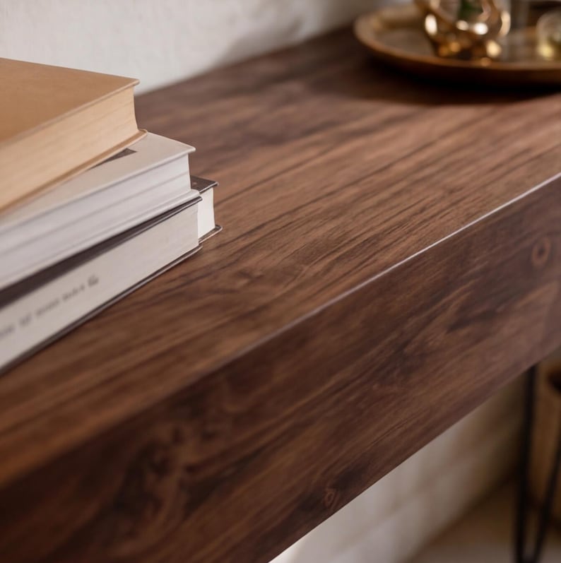 Puede incluir: Primer plano de una mesa de madera marr&oacute;n oscuro con una pila de libros en el lado izquierdo. La mesa tiene un rico patr&oacute;n de vetas de madera. Una bandeja dorada con objetos decorativos es visible en el fondo.