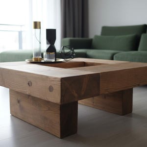 May include: A square, wooden coffee table with a recessed center. The table is constructed from thick, light brown planks and has a simple, modern design. Decorative items sit on top of the table, including a tray with glass and gold accents.