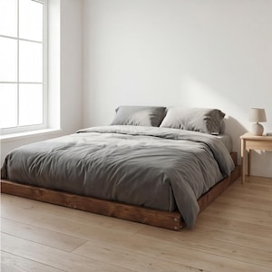 Op de afbeelding: Een bed met een laag, rustiek houten frame en een grijze dekbedovertrek en kussens. Een kleine, beige lamp staat op een houten nachtkastje. Op de achtergrond is een groot raam te zien.