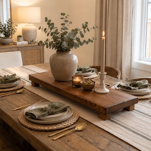 Può includere: Un tavolo da pranzo rustico in legno apparecchiato per un pasto. Il tavolo è adornato con posate, tra cui sottopiatti intrecciati, piatti bianchi e tovaglioli verdi. Un centrotavola presenta un vaso con verde, candele e un vassoio di legno.